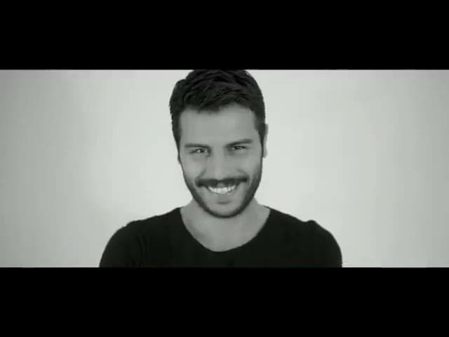 Mustachioed man in black clothing posing on a wooden chair against a brown backdrop.