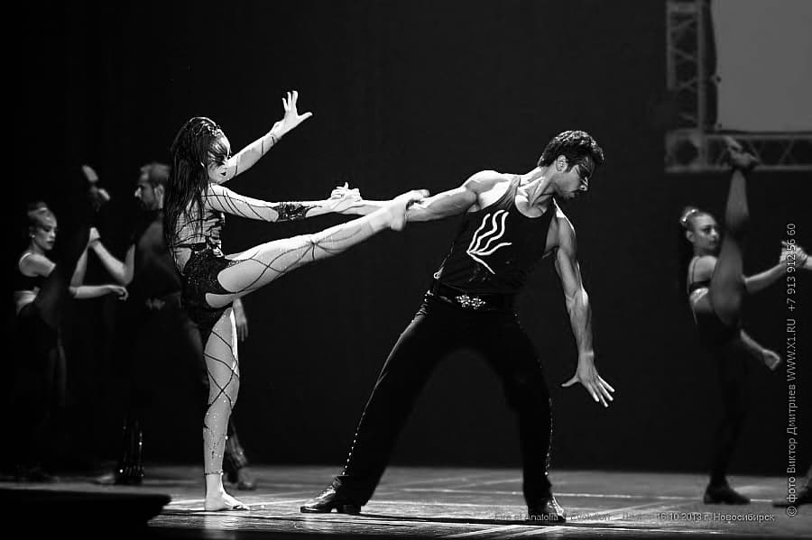 Male dancer holds female dancer's high kick in a dramatic, black and white theatrical performance.