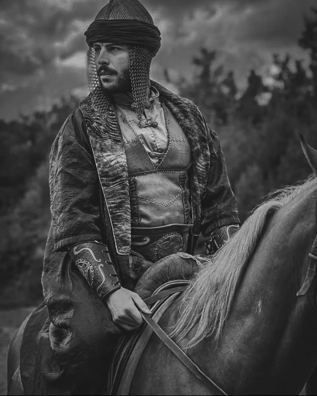 Bearded warrior in ornate historical armor, chainmail, and turban mounted on a horse in monochrome.