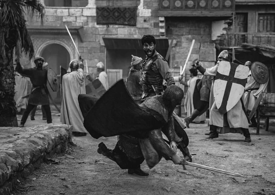 Chaotic black and white scene of medieval soldiers clashing with swords in a courtyard.