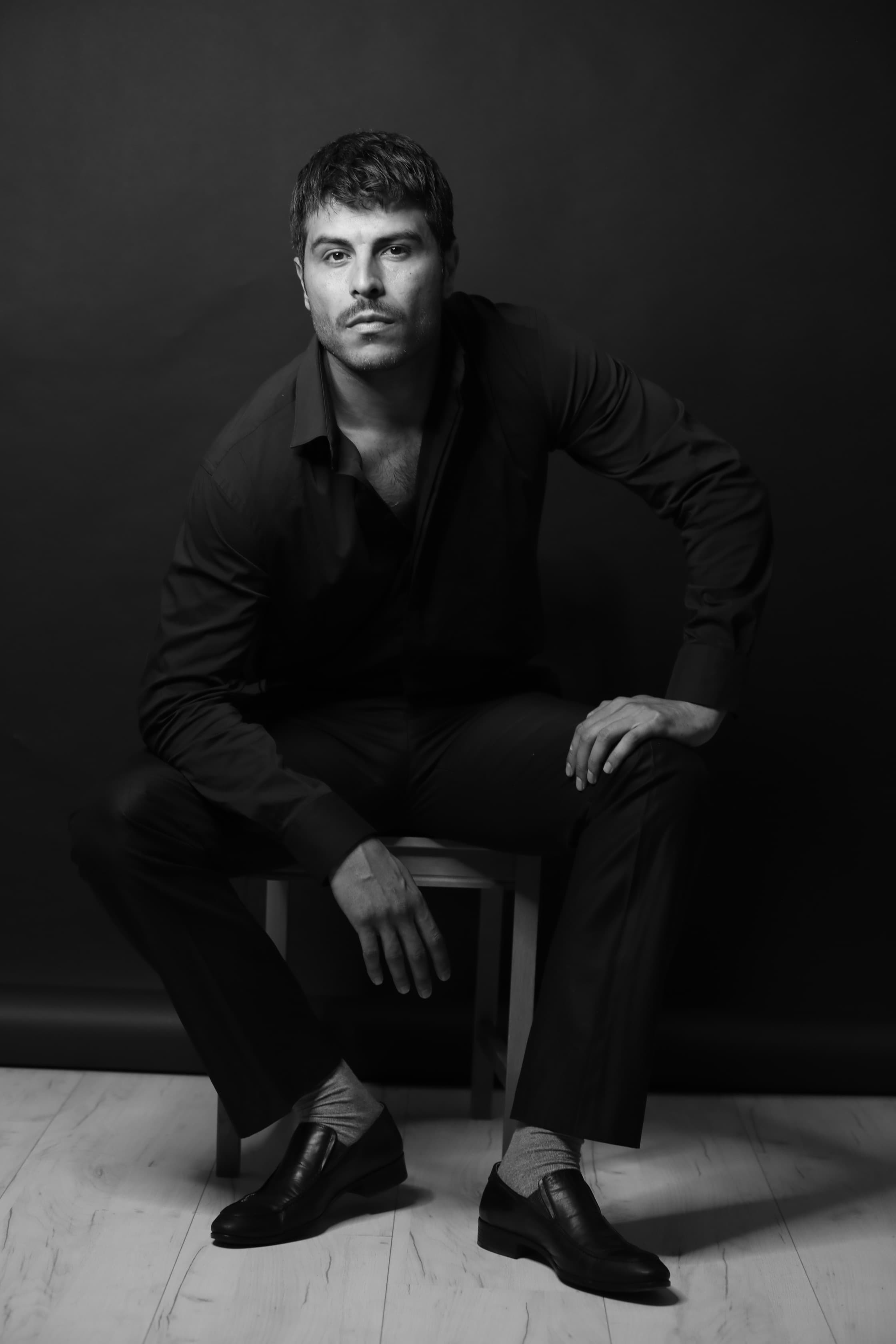 Man in dark clothing sits on a stool, leaning forward in a monochrome studio portrait.