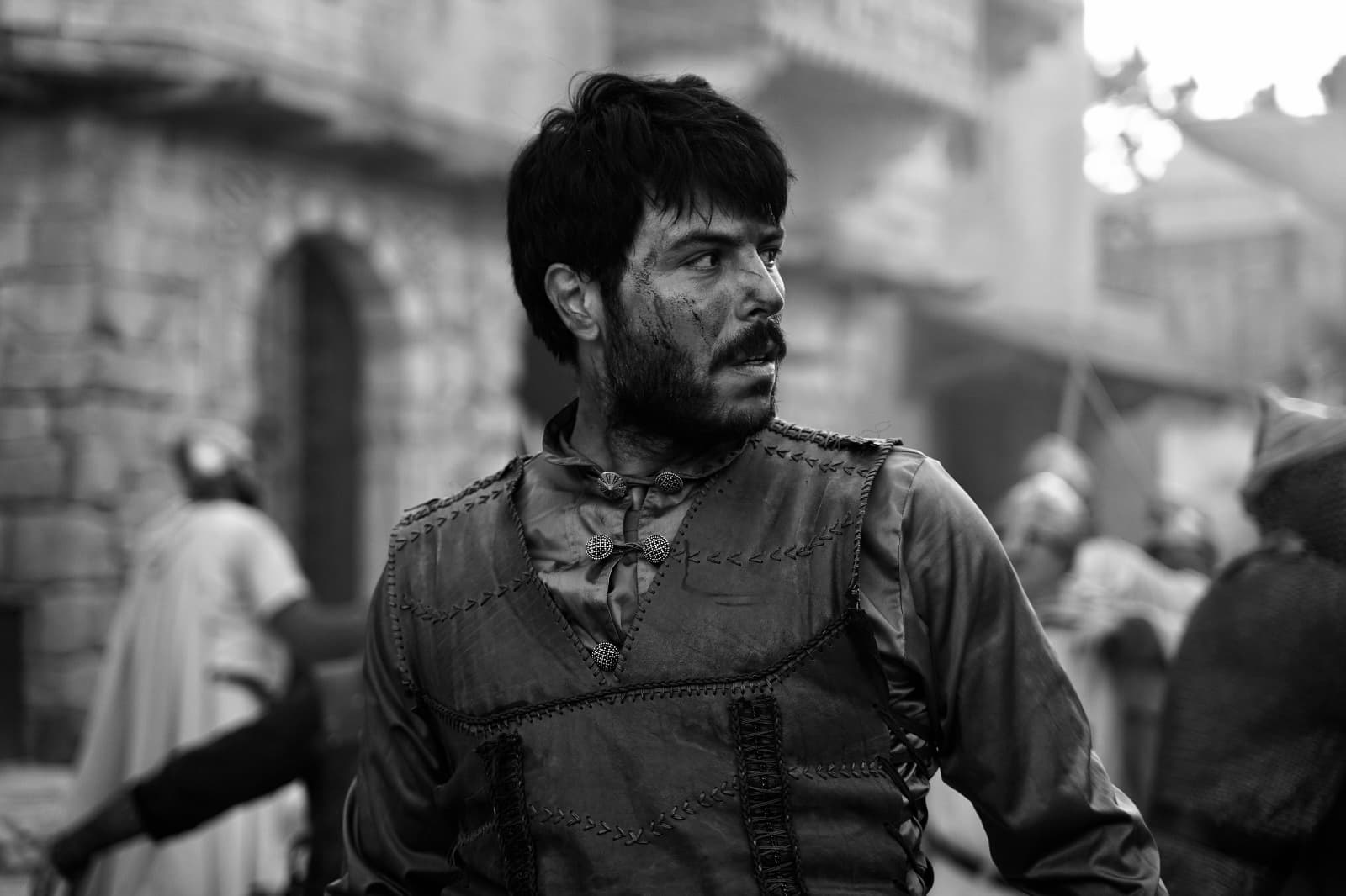 Grimy man in historical clothing looks away intensely in a black and white scene.